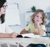 An absent-minded child staring off into space while a tutor sits by his side with an open book